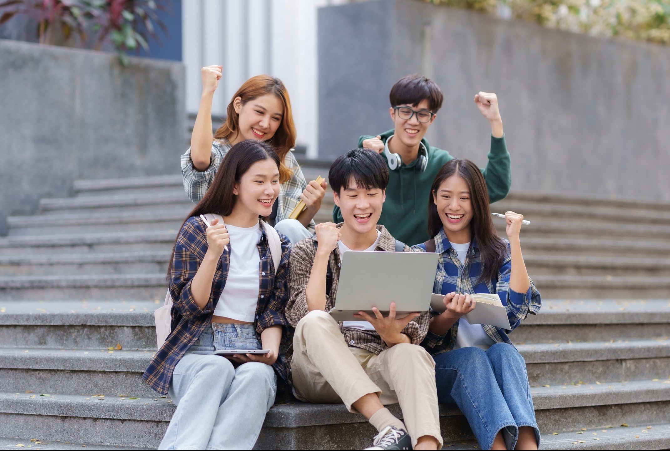 Group of Asian students with raised arms celebrate celebrating completion of exams through laptop computer.